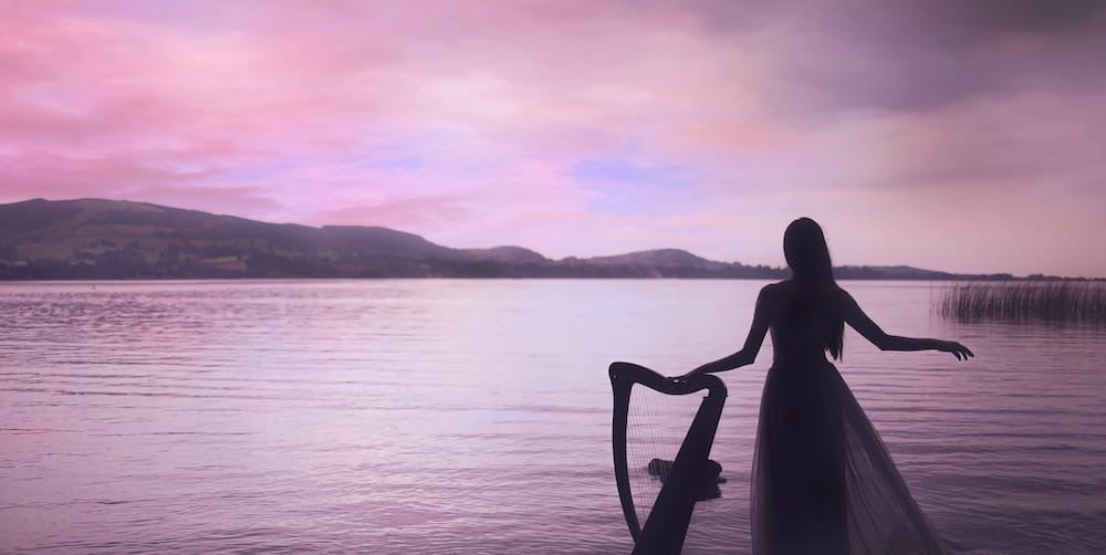 Claire Heffernan standing beside her harp, looking out over a lake.