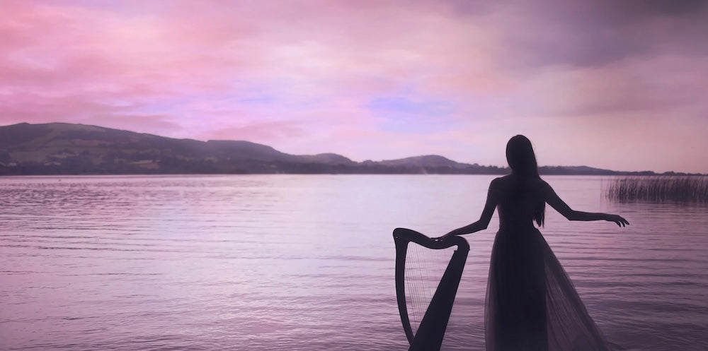 Claire Heffernan standing beside her harp, looking out over a lake.