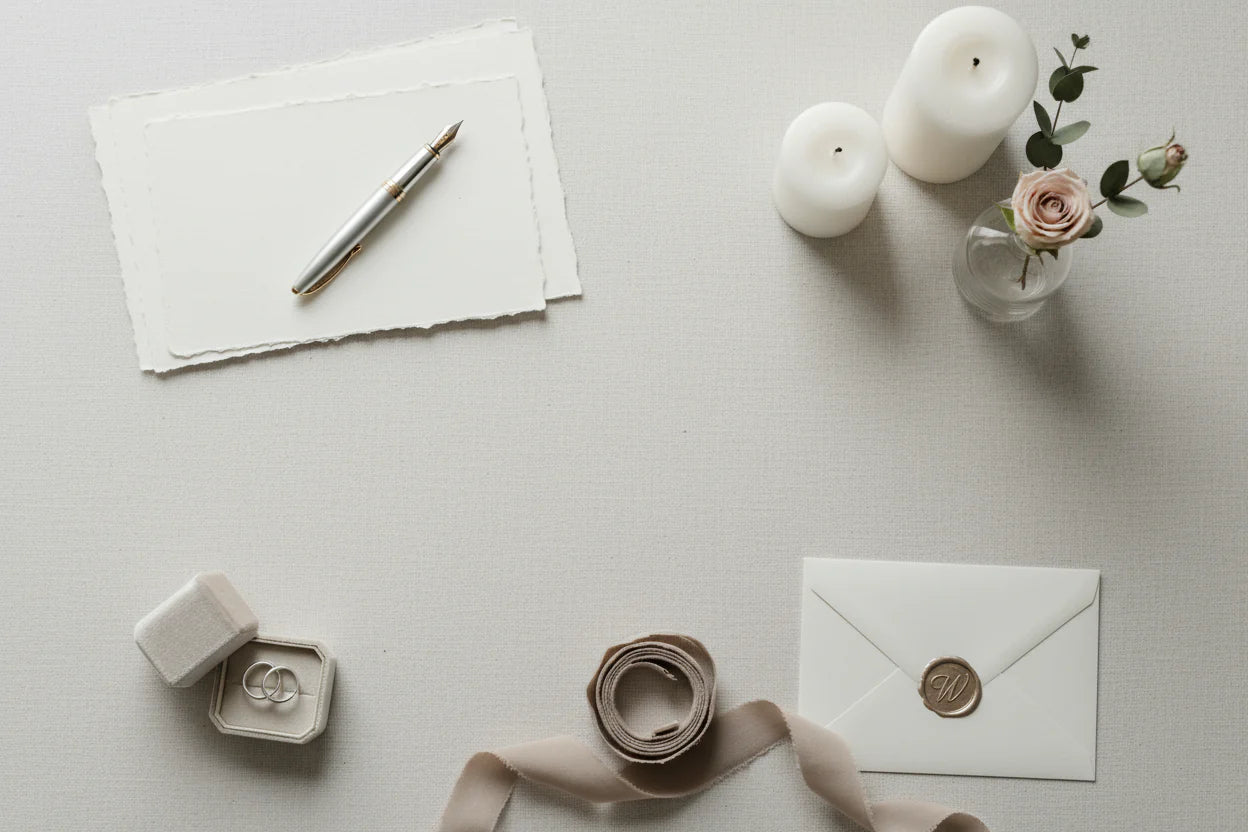 A flat lay of wedding details including a fountain pen, notepaper, a ring box, ribbon, flowers, and candles.