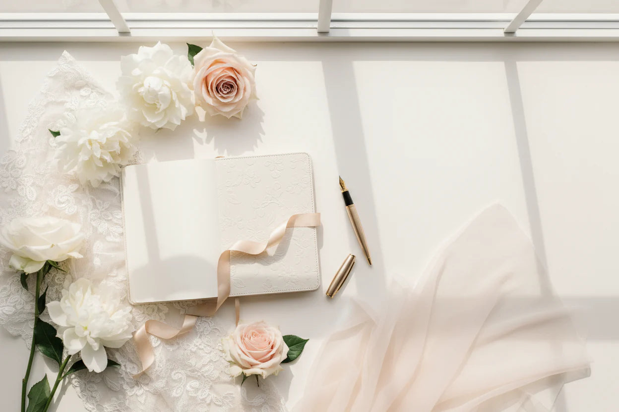 A flat lay of flowers, a notebook, and a fountain pen on a white table beside a bright window.